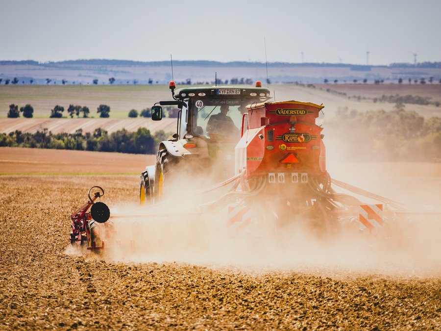 Nahaufnahme einer roten Saemaschine hinter einem Traktor, dichter Staub wirbelt ueber dem Acker