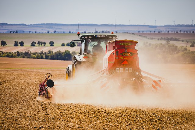 Nahaufnahme einer roten Saemaschine hinter einem Traktor, dichter Staub wirbelt ueber dem Acker