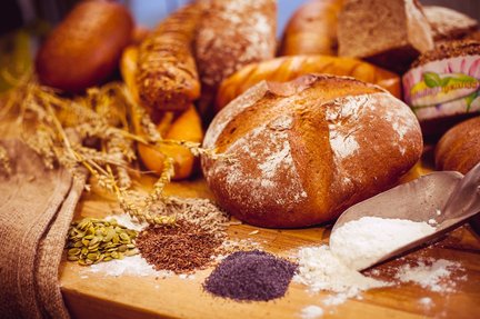Brotsortiment mit Getreide und Saaten Ein runder Brotlaib auf einem Holzbrett, davor Kuerbiskerne, Leinsamen, Mohn und Mehl, dahinter weitere Brote und Getreideaehren