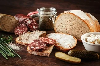 Auf einem Holztisch liegen aufgeschnittene Salami, belegte Brotscheiben, ein Laib Brot, Schnittlauch und Gewuerzgurken