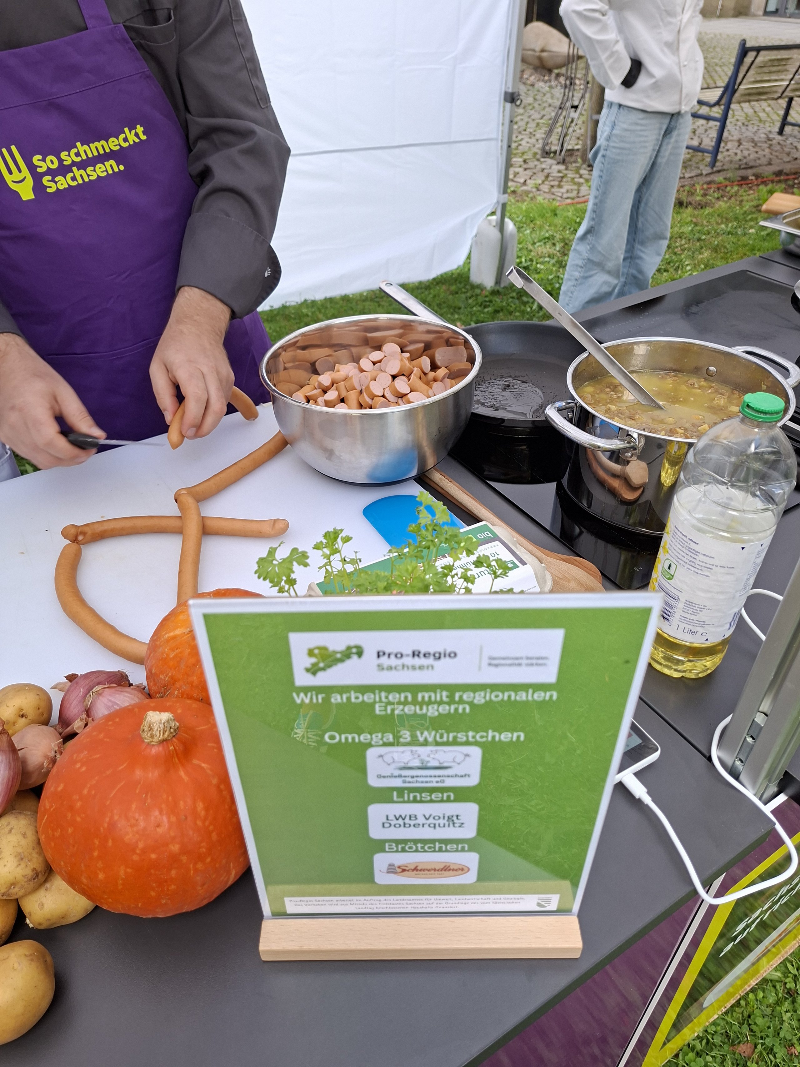 Auf einem Tisch liegen Kuerbis, Kartoffeln und Zwiebeln neben Toepfen mit Wuerstchen und Suppe, davor ein Aufsteller mit den Erzeugern