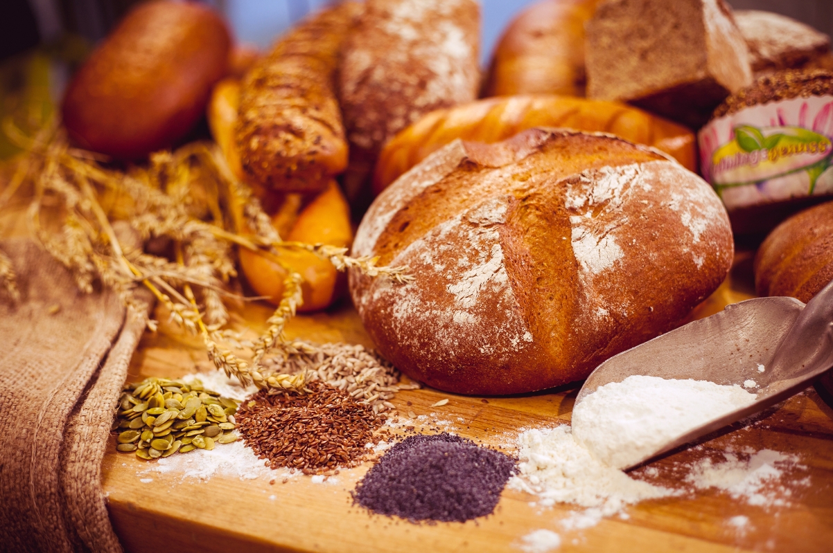 Ein runder Brotlaib auf einem Holzbrett, davor Kuerbiskerne, Leinsamen, Mohn und Mehl, dahinter weitere Brote und Getreideaehren