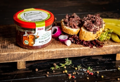 Rinder-Zwiebelmett im Glas Ein Einmachglas mit Aufstrich steht neben zwei belegten Brotscheiben auf einem Holzbrett, daneben Zwiebelringe und Gewuerzgurken