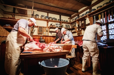 Hausschlachtung in der Stube Vier Personen in weisser Arbeitskleidung zerlegen Fleisch an einem Holztisch in einem historisch eingerichteten Raum